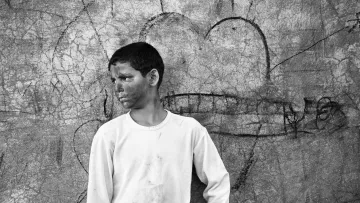 L'immagine mostra un ragazzo che sta in piedi di profilo, con un'espressione seria e pensierosa. Indossa una maglietta bianca e dei pantaloni chiari. Il suo viso appare segnato da segni di fango o polvere, il che suggerisce che potrebbe aver trascorso del tempo all'aperto o in un ambiente difficile. Dietro di lui, c'è un muro graffiato con disegni e segni, tra cui un cuore. L'atmosfera è in bianco e nero, conferendo un tono nostalgico e malinconico all'immagine.