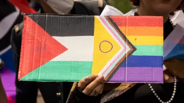 Cette image montre une personne tenant une pancarte colorée. La pancarte combine le drapeau palestinien, reconnaissable par ses bandes horizontales noire, blanche et verte avec un triangle rouge à gauche, et le drapeau Progress Pride, qui inclut les couleurs de l'arc-en-ciel ainsi que des bandes supplémentaires pour représenter la communauté transgenre et les personnes de couleur. La personne semble être en extérieur et porte une perle autour du cou ainsi que plusieurs bagues. Une autre personne portant un masque blanc est visible à l'arrière-plan.