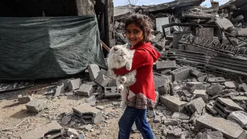 L'immagine mostra una bambina che sorride mentre tiene in braccio un coniglietto bianco. Si trova in un ambiente distrutto, con macerie e rovine visibili intorno a lei. Il suo volto esprime gioia nonostante il contesto difficile, e indossa un maglione rosso e jeans. Sullo sfondo si vedono resti di edifici e teli di plastica, che evidenziano una situazione di emergenza.