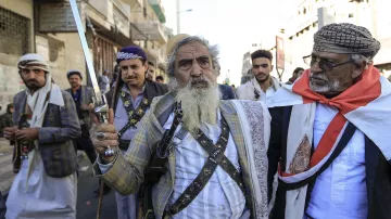 در این تصویر، چند مرد در حال راه رفتن در یک خیابان هستند. یکی از آن‌ها، که در پیش‌زمینه قرار دارد، دمی بلند و ریش سفیدی دارد و یک شمشیر در دست دارد. او لباس سنتی یمنی به تن کرده و به نظر می‌رسد که مقام یا اهمیت خاصی دارد. دیگر مردان نیز لباس‌های سنتی بر تن دارند و برخی از آن‌ها سلاح‌هایی در دست دارند. جو در تصویر به نظر مهیج و پر از نشاط است و نشان‌دهنده یک گردهمایی یا تجمع اجتماعی می‌باشد.