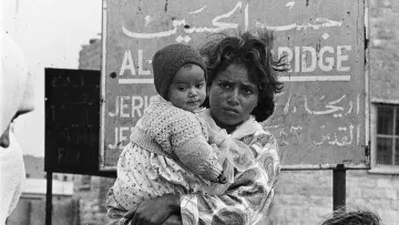 L'image montre une jeune mère, tenant dans ses bras un bébé qui porte un bonnet. La mère a un regard sérieux et semble préoccupée. En arrière-plan, on aperçoit un panneau avec des inscriptions en arabe et en anglais, indiquant des informations sur un lieu. L'atmosphère est intense, reflétant une situation de vulnérabilité. Le noir et blanc de l'image accentue l'émotion et le contexte difficile dans lequel ces personnes se trouvent.