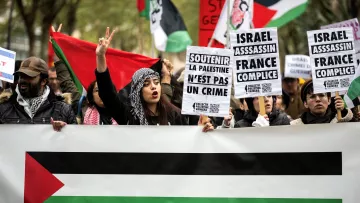 L'image montre une manifestation avec des personnes brandissant des pancartes et des drapeaux. Au premier plan, on voit une femme portant une écharpe avec un motif traditionnel qui exprime un message pacifiste en levant les deux doigts. Les pancartes contiennent des slogans concernant la Palestine, ainsi que des accusations contre Israël. À l'arrière-plan, des drapeaux aux couleurs du drapeau palestinien sont visibles, créant une atmosphère de revendication politique et de solidarité.