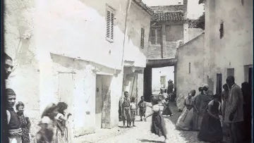 L'image représente une scène de rue dans un quartier de Constantine, en Algérie. On y voit des maisons blanches avec des volets, et une ruelle pavée qui semble étroite. Plusieurs personnes, y compris des enfants, sont visibles, certaines marchant et d'autres se tenant debout. Les vêtements des habitants semblent traditionnels, reflet d'une époque passée. L'atmosphère générale évoque une vie quotidienne animée dans un contexte historique.