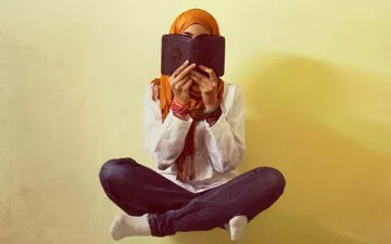 L'image montre une personne assise en tailleur, tenant un livre près de son visage, cachant ainsi son identité. Elle porte un hijab orange et une tenue simple, avec des jeans. L'arrière-plan est d'une couleur claire, créant une ambiance douce et calme. Les bras sont repliés et des bracelets colorés ornent ses poignets. Cette image évoque un moment de réflexion ou de lecture.
