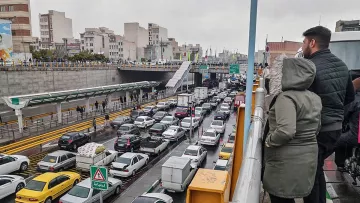 تصویر نشان‌دهنده یک صحنه شلوغ از ترافیک در یک خیابان شهری است. در سمت چپ تصویر، افرادی در حال تماشا از بالا ایستاده‌اند. در پایین، خطوط خودروها به وضوح قابل مشاهده است که نشان‌دهنده تجمع و ترافیک سنگین است. بقیه عناصر شهری مانند ساختمان‌ها و علائم راهنمایی نیز در پس‌زمینه دیده می‌شوند. هوای ابری و احتمالا بارانی به دید فضا و حس و حال تصویر اضافه می‌کند.