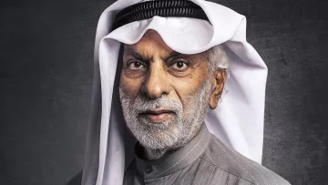 الصورة تظهر رجلاً مسنًا يرتدي الزي التقليدي العربي. لديه لحية رمادية وشعر أبيض، ويرتدي غترة بيضاء مع عقال أسود. يحمل ابتسامة لطيفة ويبدو أنه يتمتع بحضور مهيب. الخلفية مظلمة قليلاً مما يبرز تفاصيل وجهه وملابسه.