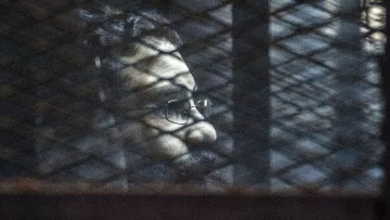 L'image montre un homme avec des lunettes et une barbe, dont le visage est partiellement obscurci par des ombres. On aperçoit des mailles ou des barres qui créent un effet de grille à l'avant, suggérant qu'il se trouve derrière une sorte de barrière ou de grillage. L'éclairage de la scène est sombre, contribuant à une ambiance mystérieuse.