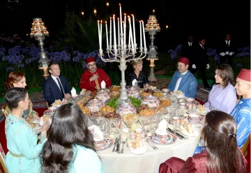 L'image montre une scène de dîner élégant, probablement dans un cadre extérieur. Une grande table est dressée avec une variété de plats et de délicieuses spécialités culinaires. De nombreuses bougies sont allumées, créant une ambiance chaleureuse et festive. Des convives, habillés en vêtements traditionnels, sont assis autour de la table, engageant des discussions animées. Au fond, on peut apercevoir des éléments décoratifs et des personnes en costume qui ajoutent à l'atmosphère formelle de l'événement.