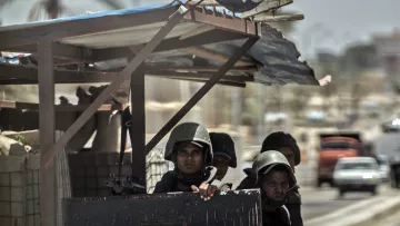 L'image montre un groupe de soldats, probablement en uniforme militaire, postés à un poste de contrôle. Ils semblent attentifs et vigilants, avec des casques de protection. On remarque également un drapeau qui flotte sur le poste, indiquant un contexte éventuellement national ou militaire. L'arrière-plan dépeint une rue, suggérant une ambiance urbaine, et on peut apercevoir des véhicules au loin. Le temps semble clair, mais la scène pourrait transmettre une sensation de tension ou de vigilance.
