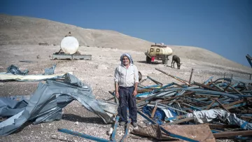 The image depicts an elderly man standing amidst a desolate landscape scattered with debris, including metal and fabric remnants. In the background, there are cylindrical tanks and a horse. The surrounding terrain features barren hills under a clear blue sky. The man appears to be wearing traditional clothing and is positioned centrally in the scene, which conveys a sense of solitude and hardship.