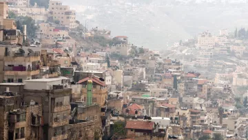 The image shows a densely populated hillside area with a mix of residential buildings. The structures vary in height and architectural style, with many appearing stacked closely together. Some roofs are visible, displaying a range of materials, from tiles to concrete. In the background, more houses and buildings can be seen on the sloping terrain, suggesting a continuation of urban development. The overall scene has a somewhat hazy atmosphere, possibly due to distance or weather conditions.