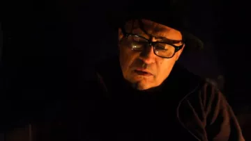 L'image montre un homme avec des lunettes et un chapeau. Il semble pensif et est plongé dans une lumière tamisée, ce qui crée une atmosphère sombre et introspective. Son expression faciale suggère qu'il est en train de réfléchir profondément ou de vivre un moment d'émotion.
