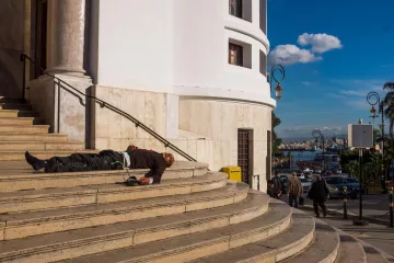 L'image montre un homme allongé sur les marches d'un bâtiment au style architectural classique, probablement en train de dormir ou en état de détresse. Le bâtiment est bien éclairé et ses murs sont de couleur claire, avec des colonnes visibles. En arrière-plan, on peut apercevoir une ville animée, avec des gens qui marchent et un port. Le ciel est bleu avec quelques nuages, ce qui donne une ambiance contrastée à la scène.