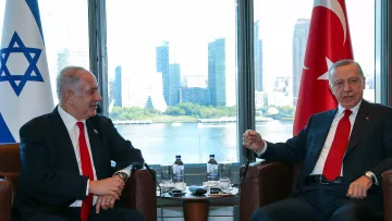 L'image montre deux hommes en discussion dans un cadre professionnel. À gauche, il y a un homme avec une cravate rouge, assis sur une chaise, regardant l'autre homme. À droite, l'autre homme, également en cravate, semble parler et gesticuler. En arrière-plan, on aperçoit des gratte-ciel et une vue sur l'eau, suggérant un environnement urbain. Des drapeaux d'Israël et de Turquie sont visibles, indiquant le contexte de cette rencontre.