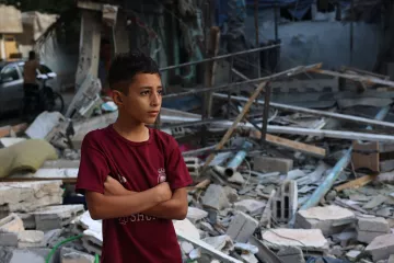 L'immagine mostra un ragazzo con le braccia incrociate, che sembra riflettere o osservare attentamente. Sullo sfondo, ci sono macerie e detriti, suggerendo che si trova in un'area devastata, probabile frutto di un conflitto o di una calamità. Il suo sguardo serio e pensieroso comunica un senso di tristezza e resilienza di fronte a una situazione difficile. La scena evidenzia anche la vulnerabilità dei bambini in contesti di crisi.