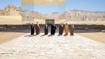 تظهر في الصورة مجموعة من الأشخاص يرتدون الزي التقليدي، ويقفوا على طريق مرصوف بالحجارة في منطقة صحراوية. خلفهم توجد جدران عاكسة تعكس المنظر الجبلي المحيط، مما يعطي شعوراً بالتواصل بين البشر والطبيعة. السماء زرقاء وصافية، مما يضيف جمالاً على المشهد.