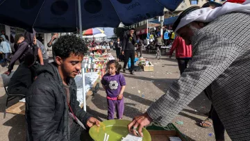 در تصویر، یک بازار محلی به چشم می‌خورد. در وسط تصویر، مرد جوانی نشسته و در حال فروش کالاهایی است. او یک بشقاب سبز رنگ را در دست دارد که در آن چند قلم وجود دارد. به نظر می‌رسد که او به مشتری‌اش توجه دارد که در حال انتخاب کالاهاست. در کنار آن، یک دختر بچه نیز در حال بازی کرده و به صحنه نگاه می‌کند. در پس‌زمینه، افراد دیگری در حال خرید و گشت و گذار هستند و جوی شلوغ و پرجنب‌وجوش به چشم می‌خورد. سایه چترهایی که بالای سر برخی افراد است، فضایی دلپذیر ایجاد کرده است.