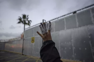 L'image montre une main levée avec des bandages enroulés autour de plusieurs doigts. En arrière-plan, on peut apercevoir un mur en métal, probablement une clôture ou une barrière, ainsi qu'un ciel nuageux. Une palme est visible à gauche, suggérant un environnement urbain. L'atmosphère semble un peu sombre et mystérieuse.