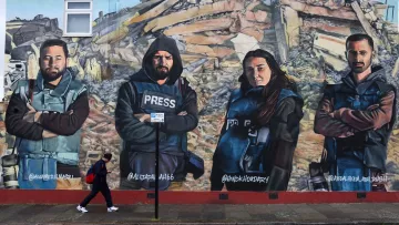 L'image montre une fresque murale représentant quatre personnes habillées en gilets pressés, posant avec les bras croisés. Ils semblent symboliser des journalistes ou des reporters. En arrière-plan, il y a des débris, suggérant un contexte de destruction ou de conflit. Une personne marche au premier plan, ajoutant une dynamique à la scène. Les visages des individus sur la fresque sont sérieux et déterminés.