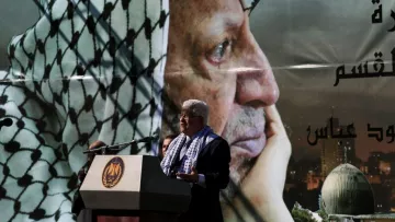 L'image représente un homme en train de parler lors d'un discours. Il se tient devant un grand poster qui affiche le portrait d'un homme en keffieh, regardant pensivement au loin. Le décor suggère qu'il s'agit d'un événement commémoratif, probablement lié à des thèmes politiques ou historiques. Le podium sur lequel l'homme s'exprime est orné d'un emblème officiel, tandis que le fond montre une ville, ajoutant un aspect symbolique à la scène.