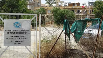 L'image montre une zone entourée d'une clôture. À gauche, il y a un panneau de la zone tampon des Nations Unies, indiquant qu'il s'agit d'un espace où l'entrée est interdite sans autorisation. À l'arrière-plan, on peut voir des bâtiments avec des antennes sur le toit, ainsi que de la végétation désordonnée au sol, créant une atmosphère de négligence. Le tout semble évoquer une région où l'accès est strictement contrôlé, probablement en raison de tensions historiques.