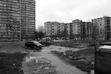 L'image montre un paysage urbain qui semble désolé. On peut voir plusieurs immeubles de grande hauteur, typiques de l'architecture des banlieues. Le sol est quelques peu inégal, avec des flaques d'eau et des débris éparpillés, ce qui donne une impression de négligence. Deux voitures sont garées sur un chemin de terre. L'environnement est sombre, accentué par le ciel nuageux, ce qui ajoute à l'atmosphère générale de tristesse et de délabrement.