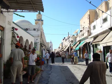L'image montre une rue animée d'une ville typique, avec des bâtiments de style méditerranéen. On peut voir des personnes marchant le long de la rue, parmi lesquelles des hommes habillés en vêtements traditionnels. Des pots de fleurs et des végétaux décorent l'entrée de certains bâtiments, tandis que des ombrelles ou des auvents semblent offrir de l'ombre aux passants. Le ciel est dégagé et ensoleillé, créant une ambiance agréable.
