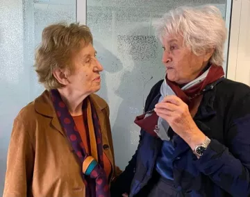 L'image montre deux femmes âgées engagées dans une conversation. Elles semblent amicales et souriantes. L'une porte une veste marron avec un foulard à pois, tandis que l'autre est habillée de manière similaire, avec une écharpe. L'arrière-plan est flou, mettant en valeur leur interaction.
