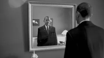 L'image montre un homme en costume noir se tenant devant un miroir. Son reflet dans le miroir affiche une expression sérieuse. L'environnement semble sobre, avec des éléments de décoration tels qu'une lampe et un cadre au mur. La scène est présentée en noir et blanc, ajoutant une atmosphère mélancolique et introspective à l'image.