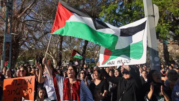 L'image montre une manifestation où des personnes tiennent des drapeaux palestiniens et des pancartes. La foule semble rassemblée pour exprimer une opinion ou un soutien, avec des femmes et des enfants présents. L'arrière-plan inclut des arbres et un environnement urbain. Les participants portent des vêtements variés, certains ayant des motifs traditionnels. L'atmosphère paraît chargée d'émotion et de solidarité.