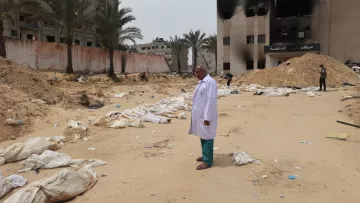 L'immagine mostra un paesaggio desolato e drammatico, caratterizzato da un terreno sabbioso. Al centro, un uomo in un camice bianco osserva un'area circondata da edifici danneggiati, con alcuni segni di distruzione visibili sullo sfondo. Intorno a lui ci sono sacchi bianchi disposti a terra, che suggeriscono una scena di grande sofferenza e perdita. Palme si ergono sullo sfondo, ma l'atmosfera generale è segnata da un senso di tristezza e desolazione.