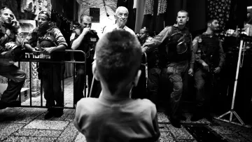 L'image montre un enfant vu de l'arrière, regardant vers un groupe de personnes, dont certaines semblent être des policiers ou des soldats. L'atmosphère est tendue, avec des barrières de sécurité en place et une composition en noir et blanc qui accentue le contraste entre les figures. L'enfant, central dans la scène, semble intrigué ou préoccupé par la situation qui se déroule devant lui. Les autres personnes sont alignées, probablement en position d'intervention.