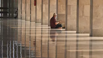 L'image montre un homme assis sur le sol d'un espace intérieur, probablement un grand hall ou une mosquée, avec des murs de pierre ainsi qu'un sol brillant et réfléchissant. L'homme porte un chapeau et lit un livre, et l'environnement est calme et serein. Il semble être plongé dans sa lecture, tandis que la lumière naturelle crée des reflets sur le sol.