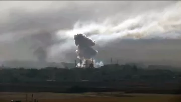 La imagen muestra una escena con humo denso y nubes oscuras que se elevan desde un área urbana. Se aprecian instalaciones industriales o edificios en el fondo, y el humo parece provenir de una explosión o un incendio. El cielo está nublado, lo que contribuye a una atmósfera sombría y tensa en la imagen.