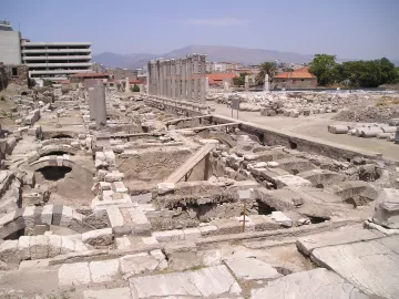 L'image montre un site archéologique avec des ruines et des structures anciennes. On aperçoit des colonnes dressées, probablement d'un bâtiment antique, ainsi que des fondations de murs en pierre. En arrière-plan, on peut distinguer des bâtiments modernes, contrastant avec les vestiges historiques. Le ciel est clair et le paysage est légèrement vallonné au loin. L'endroit semble être une zone de fouilles, avec des éléments qui témoignent d'une ancienne civilisation.