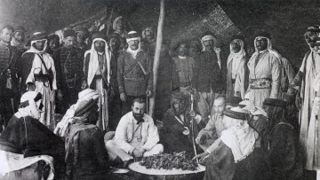 این تصویر تاریخی گروهی از مردان را در یک محیط سنتی نشان می‌دهد. آنها در یک چادر یا فضای باز گرد هم آمده‌اند و به نظر می‌رسد در حال تهیه یا صرف غذا هستند. چند نفر از آن‌ها لباس‌های سنتی عربی به تن دارند، از جمله عبا و headscarf (کشیه). در مرکز تصویر، چند نفر در حال نشستن و گفتگو هستند و سایرین ایستاده یا نشسته در اطراف آن‌ها قرار دارند. فضای تصویر گرم و صمیمی به نظر می‌رسد و نمایانگر فرهنگی غنی و سنت‌های اجتماعی است.