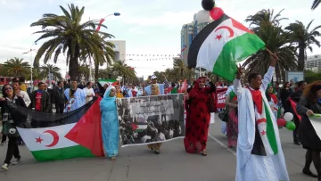 The image depicts a lively demonstration, likely in support of a cause related to Western Sahara. People are marching along a street, holding banners and flags that feature the colors red, green, and black, which are associated with the Sahrawi nationalist movement. The crowd includes both men and women, some wearing traditional clothing. In the background, palm trees and buildings suggest a warm climate, and festive decorations, like balloons, add to the atmosphere of the event. The banner appears to convey a message related to their cause.