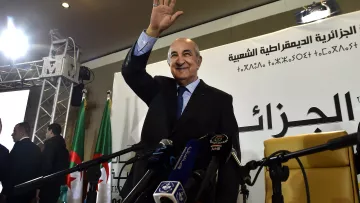 L'image montre un homme qui semble saluer une audience, debout devant un podium. Il porte un costume et a une main levée en signe de salut. En arrière-plan, on peut apercevoir des drapeaux, possiblement de l'Algérie, et des micros disposés devant lui, ce qui indique qu'il s'agit d'un événement officiel ou d'une conférence de presse. Des personnes semblent également présentes, apportant une ambiance formelle à la scène.