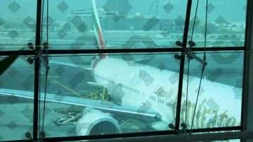 La imagen muestra un avión de la aerolínea Emirates estacionado en un aeropuerto. Se puede ver parte del fuselaje y el ala del avión a través de un gran ventanal con un diseño en las ventanas. Hay trabajadores en la plataforma que parecen estar realizando tareas de mantenimiento o preparación, y se puede observar un entorno aeroportuario con otros edificios y maquinaria en el fondo. La atmósfera es luminosa, aunque un poco difusa debido al cristal.