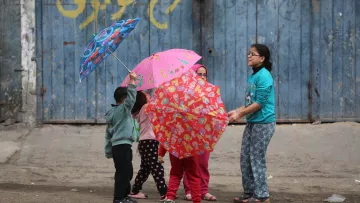 L'immagine mostra un gruppo di bambini che giocano insieme in strada, mentre tengono in mano degli ombrelli colorati. I bambini indossano abiti semplici e variopinti, e sembrano divertirsi sotto un muro blu con scritte. La scena trasmette gioia e convivialità.