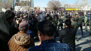 L'image montre une foule nombreuse rassemblée dans une rue, probablement en train de manifester. On observe des personnes de différents âges et sexes, certaines prenant des photos ou filmant avec leurs téléphones. Au centre de la scène, il semble y avoir un affrontement ou une tension, avec des forces de sécurité en uniforme. La situation paraît dynamique, avec des expressions faciales variées qui indiquent des émotions fortes parmi les manifestants. Des bâtiments et des enseignes sont visibles en arrière-plan, contribuant à l'ambiance de l'événement.
