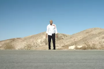 L'image montre un homme debout au milieu d'une route désertique, avec des collines arides en arrière-plan. Il porte une chemise blanche qui semble tâchée de sang et un pantalon noir. Le ciel est clair et bleu, ce qui souligne l'ambiance isolée et dramatique de la scène. Les éléments autour de lui donnent une impression de solitude et de tension.