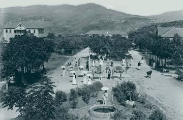 L'image semble représenter une scène rurale d'une époque passée. On peut voir un chemin central bordé d'arbres et de bâtiments à l'arrière-plan. Plusieurs personnes, vêtues de manière traditionnelle, se déplacent, certaines portant des parapluies. Des animaux comme des bœufs sont également présents, ajoutant à l'atmosphère agricole de la scène. Au premier plan, il y a un puits, probablement utilisé par les villageois. Les montagnes en arrière-plan complètent le paysage verdoyant. Cette image évoque une vie paisible et communautaire en harmonie avec la nature.
