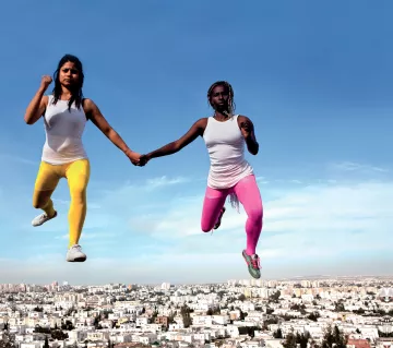 L'image montre deux femmes qui semblent flotter dans les airs, se tenant par la main. Elles portent des vêtements de sport, l'une en leggings jaunes et l'autre en leggings roses, avec des t-shirts blancs. En arrière-plan, on peut voir une ville urbaine s'étendre sous elles, avec un ciel bleu clair. Cette composition crée une sensation de liberté et de mouvement dynamique.