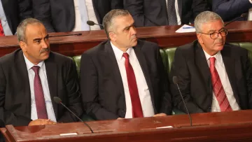 L'image montre trois hommes assis côte à côte dans un cadre formel, probablement lors d'une réunion ou d'une séance parlementaire. Ils portent des costumes sombres et des cravates, affichant une expression sérieuse. L'un d'eux porte une cravate rouge, tandis que les autres optent pour des nuances plus sombres. En arrière-plan, on aperçoit des personnes en costume, suggérant un environnement officiel.