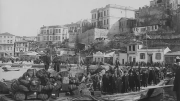 Cette image en noir et blanc montre une scène portuaire historique. On peut voir une barge chargée de marchandises, notamment des fûts et des sacs, avec des personnes debout dessus. En arrière-plan, il y a des bâtiments qui semblent faire partie d'une ville côtière, avec des structures en pierre et des maisons aux balcons. La côte est animée, avec d'autres bateaux et des gens sur le rivage, créant une atmosphère de commerce maritime. La lumière et le contraste évoquent une ambiance d'époque.