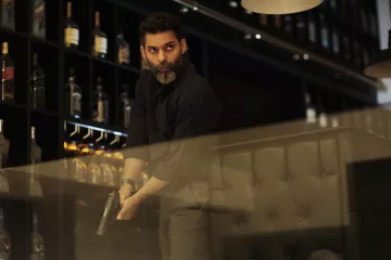 L'image montre un homme, probablement dans un bar ou un restaurant, tenant une arme à feu. Il a une barbe grise et une expression sérieuse, suggérant qu'il est dans une situation tendue. Le décor en arrière-plan comprend des étagères avec des bouteilles de spiritueux. L'éclairage est tamisé, créant une atmosphère dramatique.