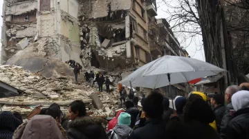 L'image montre une scène de destruction après un tremblement de terre. On peut voir un bâtiment gravement endommagé, avec des murs effondrés et des débris éparpillés. Au premier plan, une foule de personnes semble se rassembler, regardant les secours qui travaillent. Certains porteurs d'ombrelles se protègent de la pluie. L'atmosphère est sinistre, et l'on ressent une forte émotion face à la destruction causée par la catastrophe.