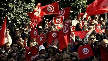 L'image montre une foule de personnes rassemblées, agitant des drapeaux tunisiens rouges avec un cercle blanc et une étoile à cinq branches. Les manifestants semblent animés, levant les bras en signe de solidarité ou de protestation. On peut voir un paysage urbain en arrière-plan avec des arbres, ce qui suggère un rassemblement en plein air. L'atmosphère semble énergique et engagée, reflétant un moment de mobilisation collective.