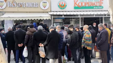 الصورة تظهر مجموعة من الأشخاص الواقفين في طابور أمام متجر يُظهر لافتة تدل على أنه نقطة بيع للمنتجات. يبدو أن معظم الأشخاص يرتدون ملابس شتوية وأنهم يحملون أكياسًا. الجو يبدو هادئًا، مع وجود أشجار حول المكان، بينما يبدو أن بعضها قد فقد أوراقه بسبب البرد. المتجر يحمل اسم "كوoperative" ويبدو أنه يبيع مواد غذائية أو منتجات محلية.